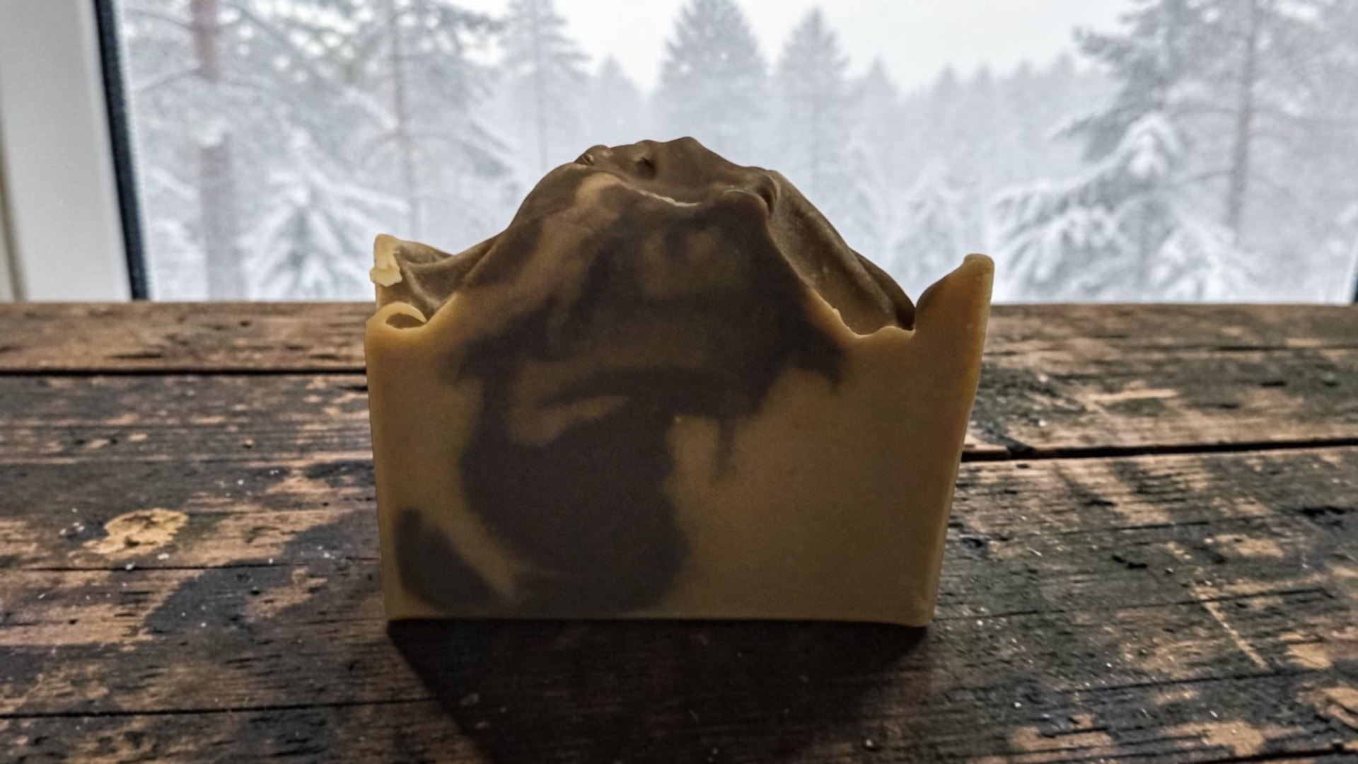 Winter In Soap, a handcrafted bar with creamy swirls on weathered wood in front of a snowy forest window