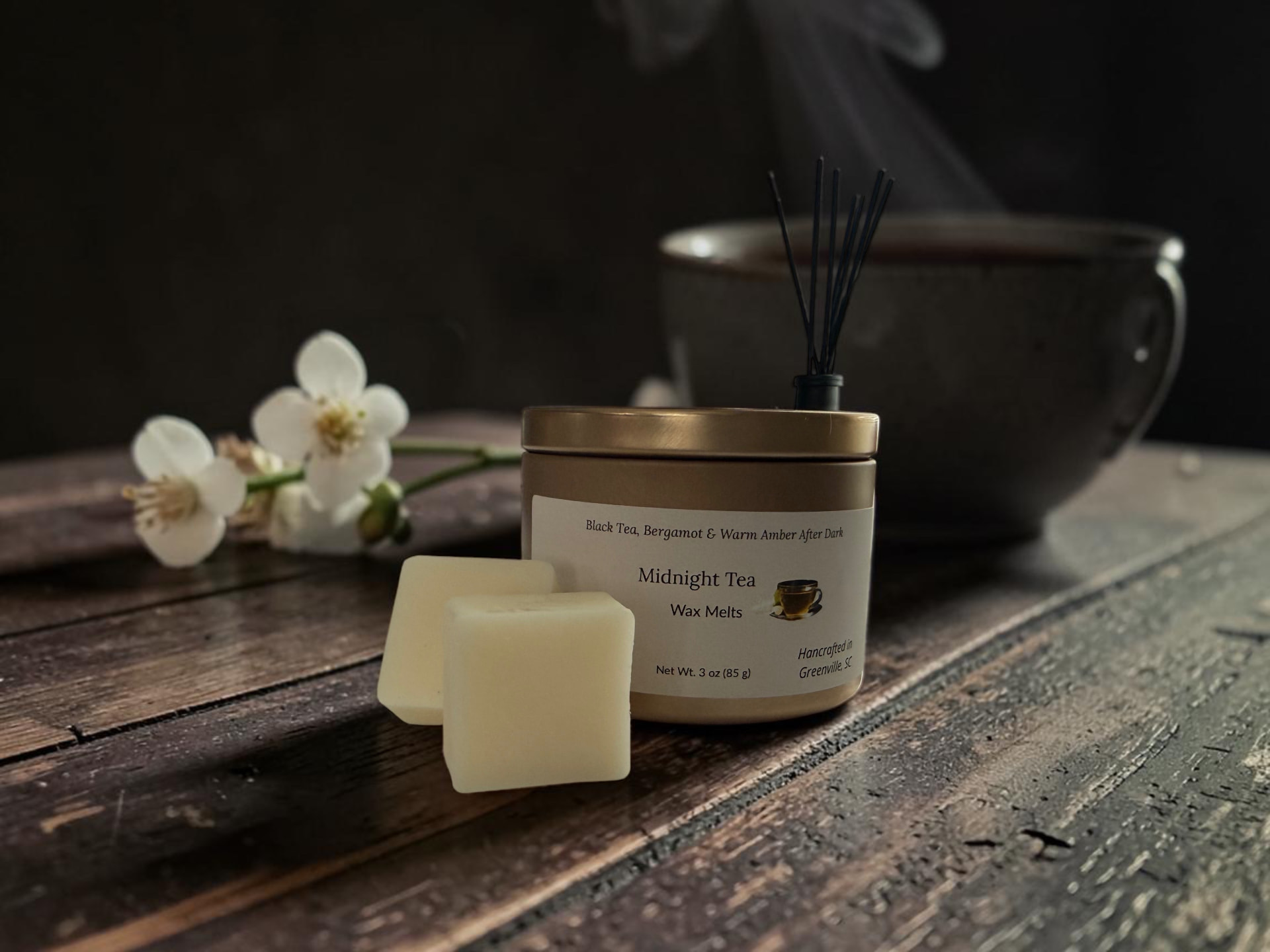 Midnight Tea Wax Melts, a gold tin with creamy wax melt squares beside white jasmine and black tea leaves on dark wood