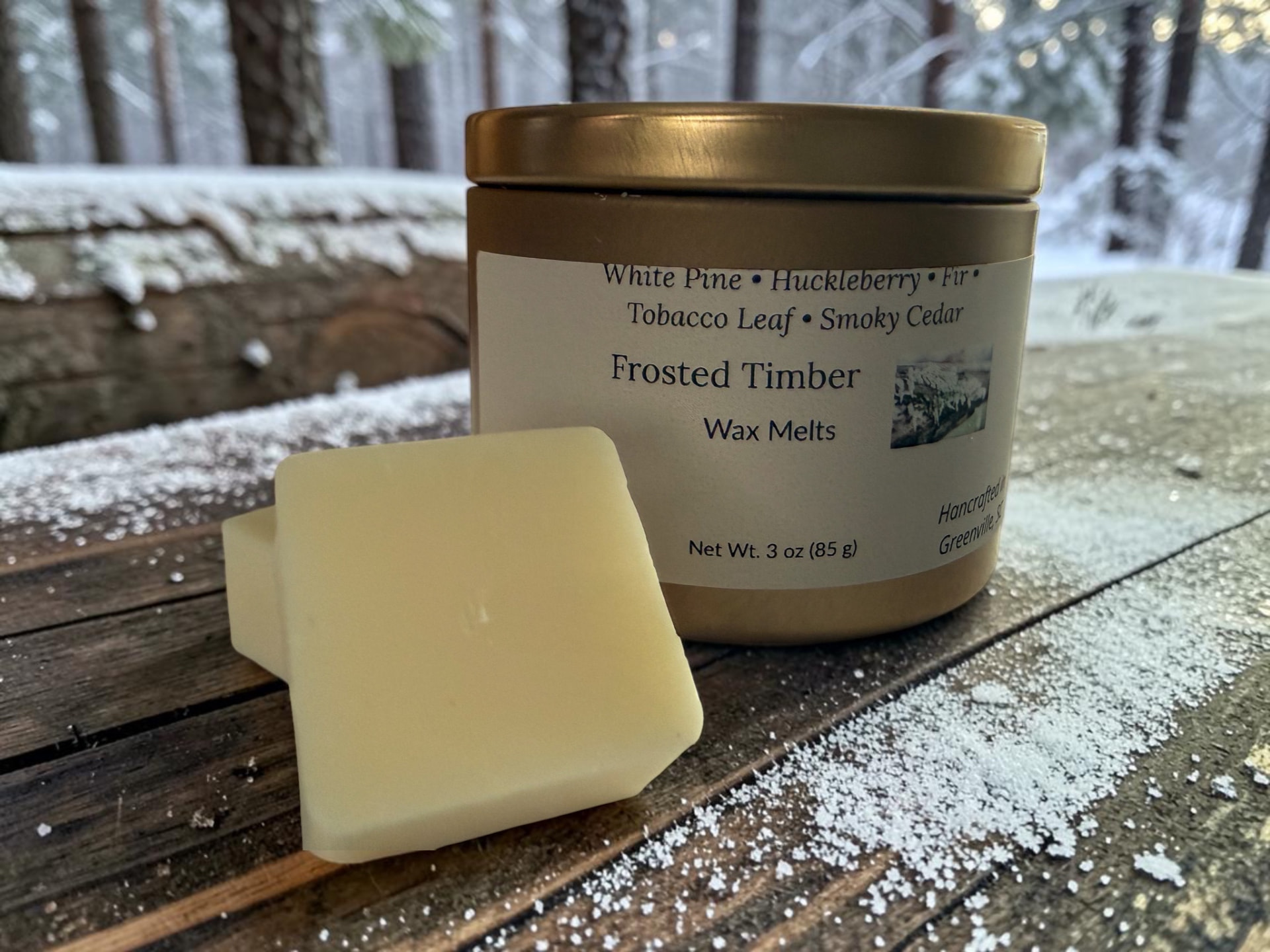 Frosted Timber Wax Melts, a gold tin with creamy wax melt squares on weathered wood with a snowy forest in the background