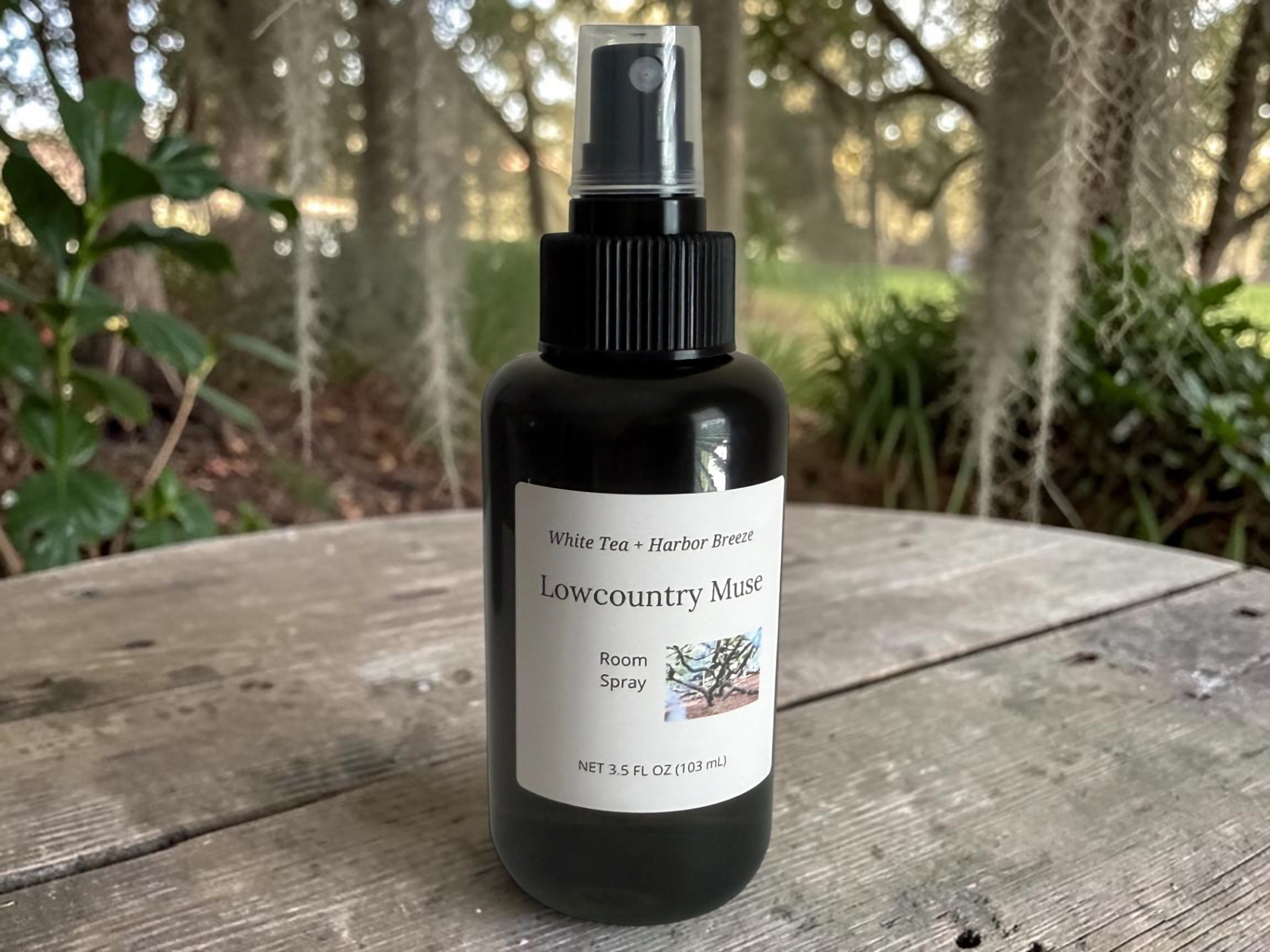 Lowcountry Muse Room Spray, a black bottle with a black spray cap on a weathered wooden surface with Spanish moss and greenery in the background